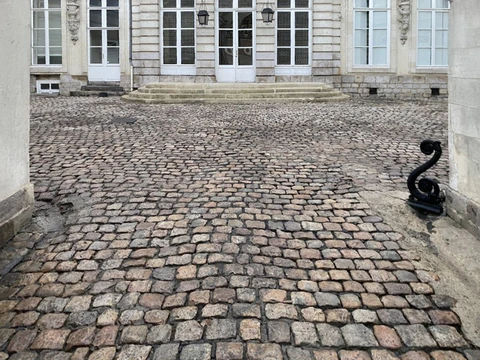 Cour pavée d'un château ou bâtiment historique français avec façade en pierre claire, portes-fenêtres à croisillons blancs et lanternes suspendues